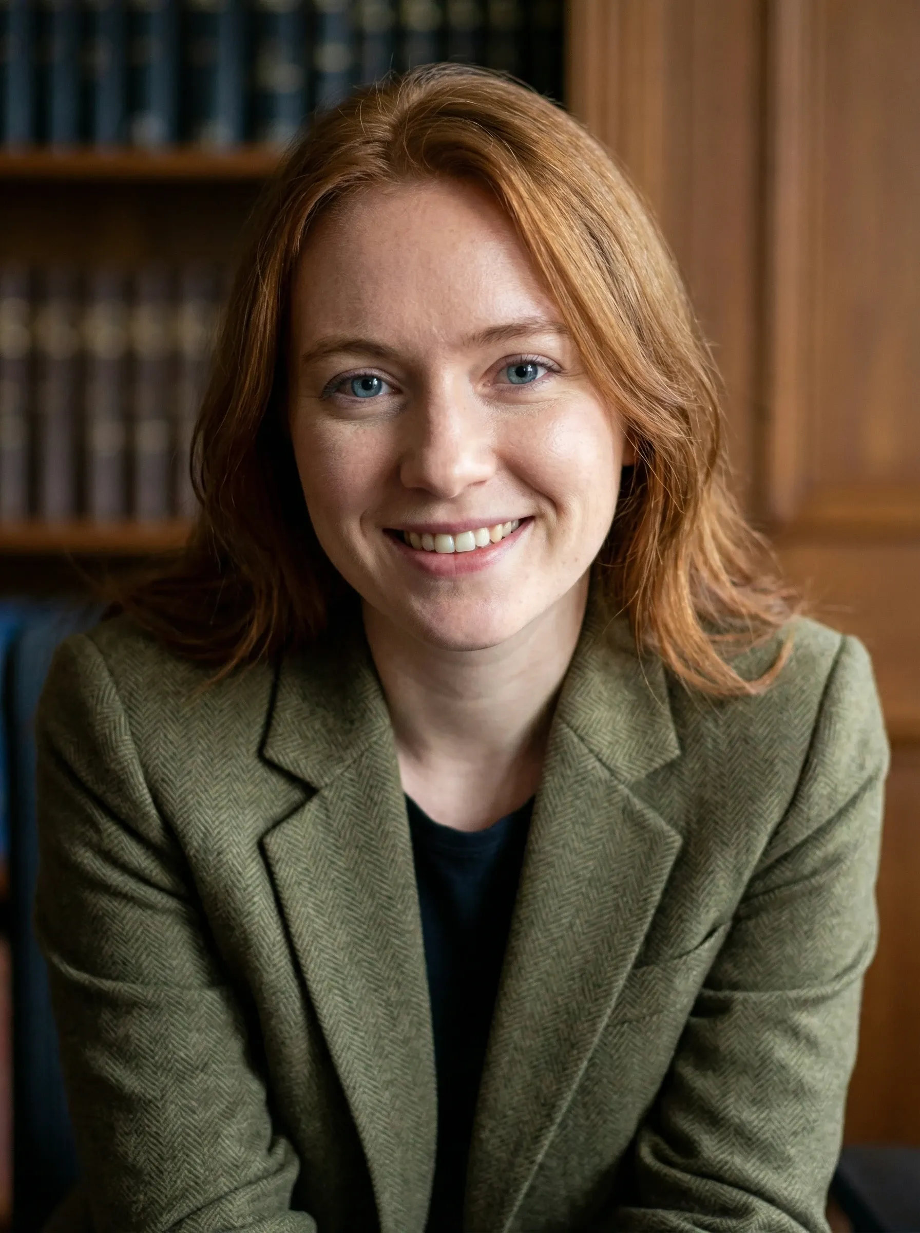 Professional headshot of a woman in an olive blazer and black top in a library setting
